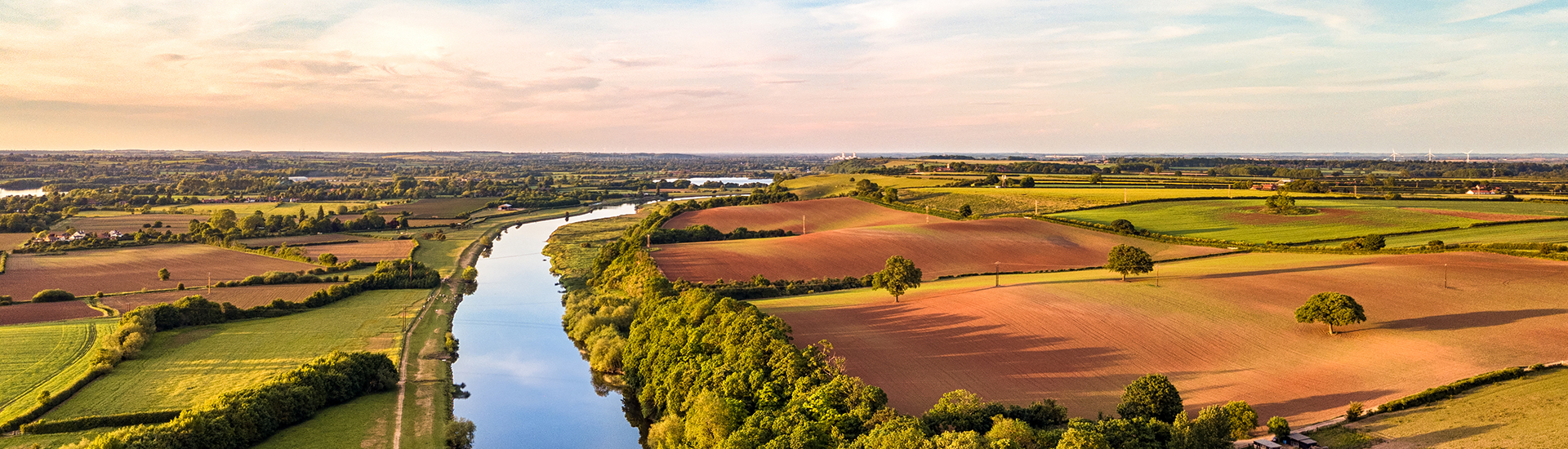 Aerial view of a river winding through a patchwork of green pastures and plowed agricultural fields at golden hour, with trees lining the riverbanks and a wide rural landscape stretching to the horizon.